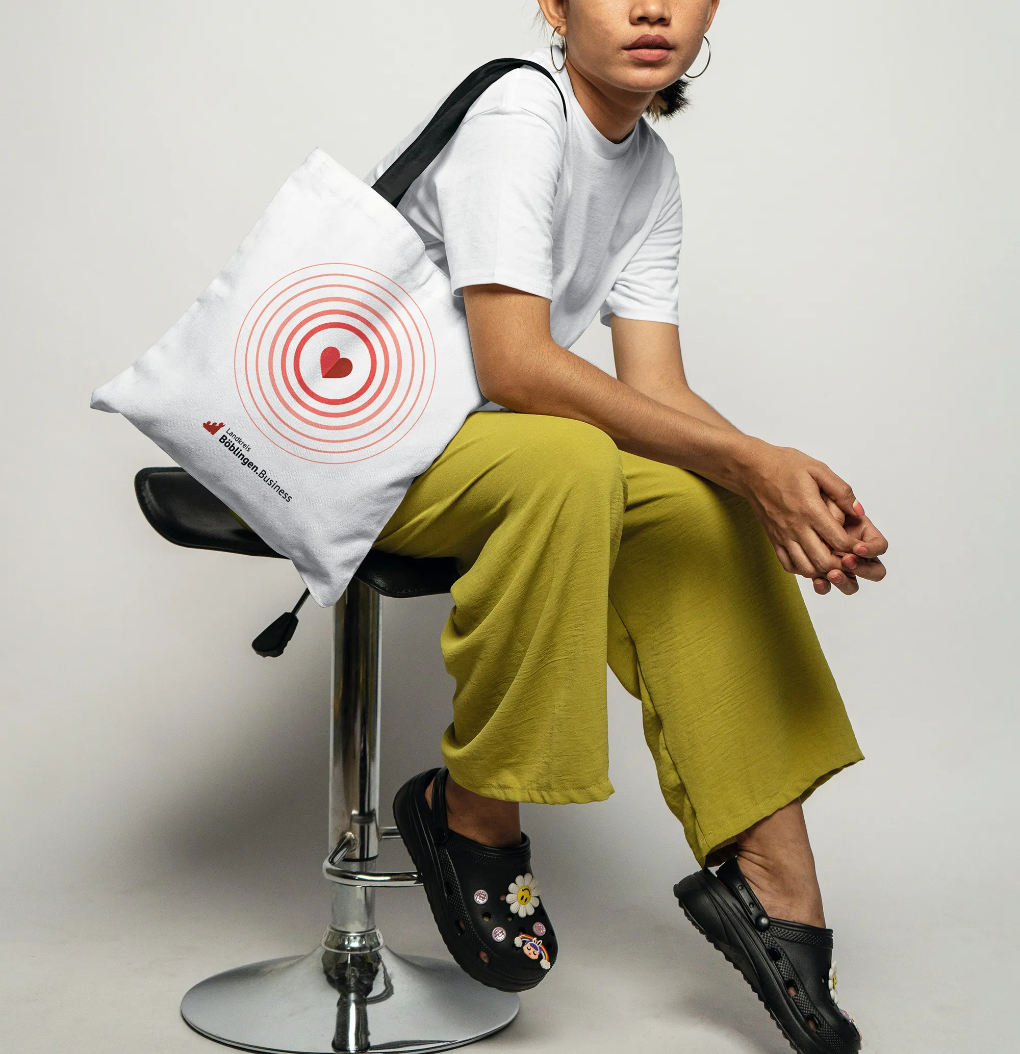 Person sitzt auf einem verchromten Barhocker mit weißer Tasche, die rote Zielscheiben und den Text ‚Landkreis Böblingen.Business‘ zeigt.