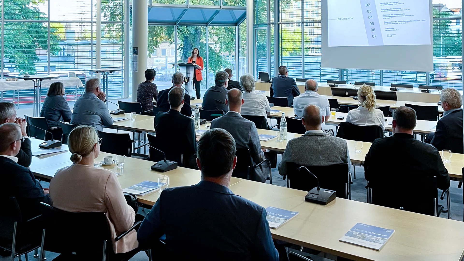 Frau hält eine Präsentation vor einem Publikum in einem modernen, hellen Konferenzraum mit großen Fenstern.