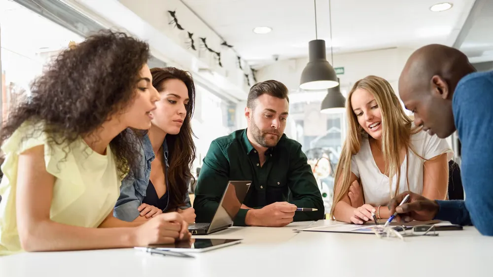 Five diverse professionals collaborating around a white table in modern office with laptops and tablets