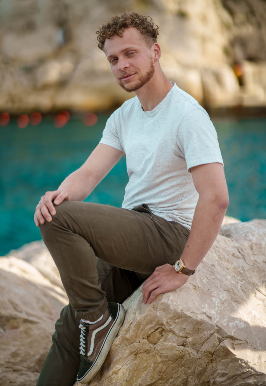 Homme assis sur un rocher près de l'eau turquoise avec un t-shirt blanc et un pantalon marron.
