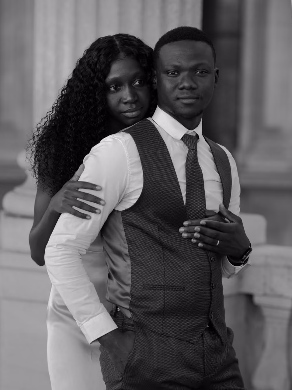 Un couple noir élégant dans une pose tendre, la femme enlace l'homme par derrière, tous deux habillés formellement.