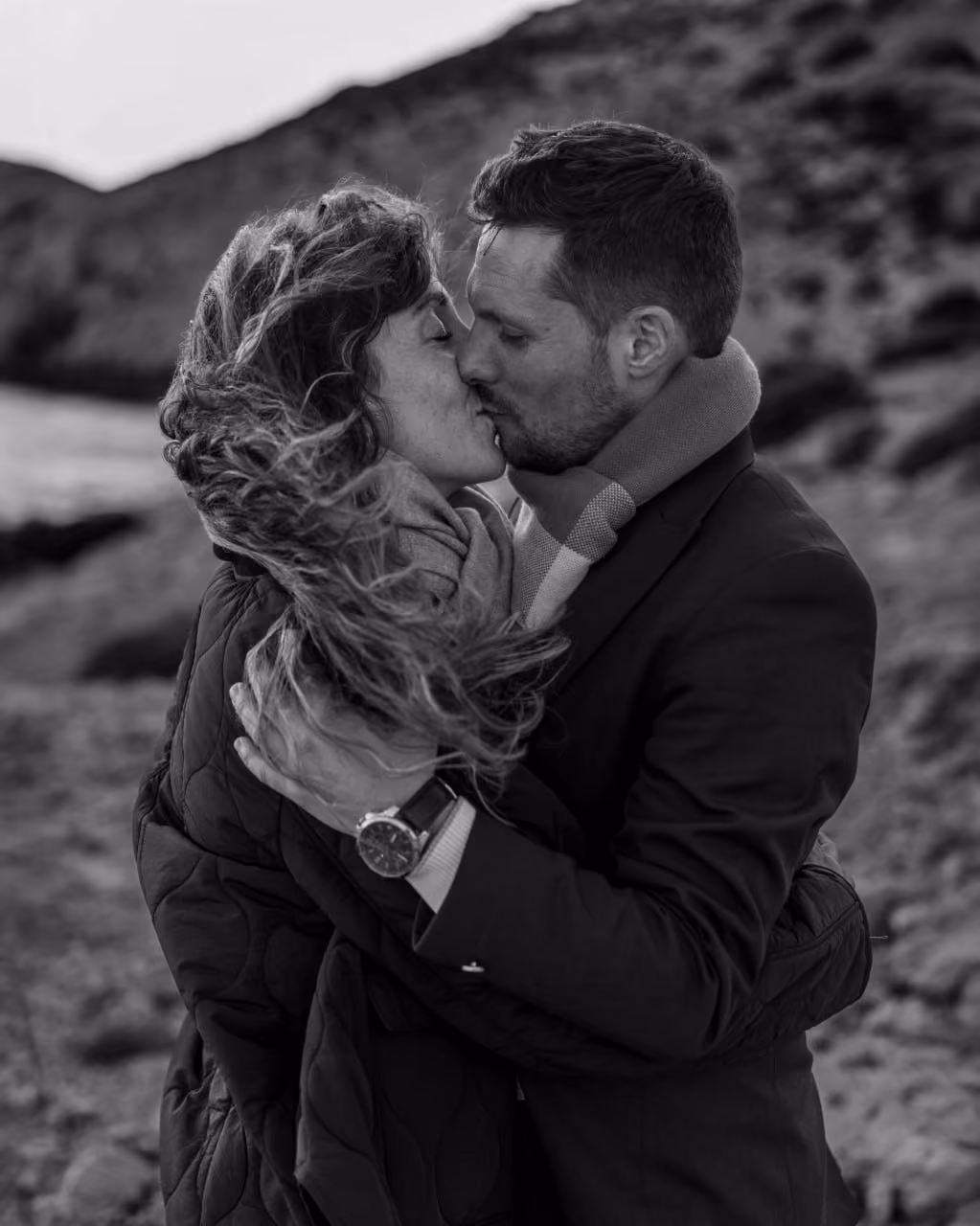 Un couple s'embrassant tendrement en extérieur avec la femme enveloppée dans une écharpe et le vent soufflant dans ses cheveux.