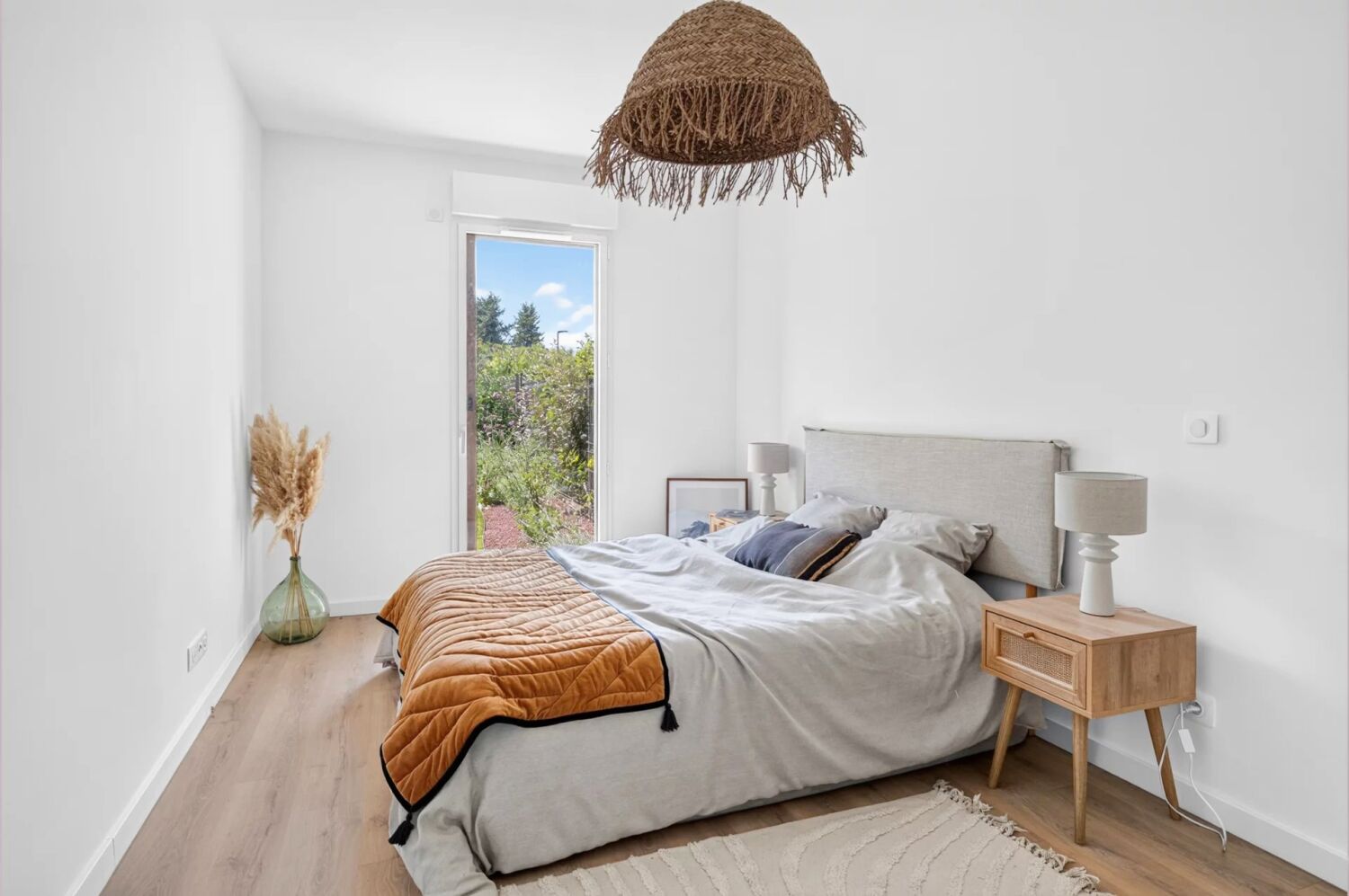 Chambre lumineuse avec lit, couverture orange, lampe, table de chevet en bois et porte-fenêtre donnant sur jardin.