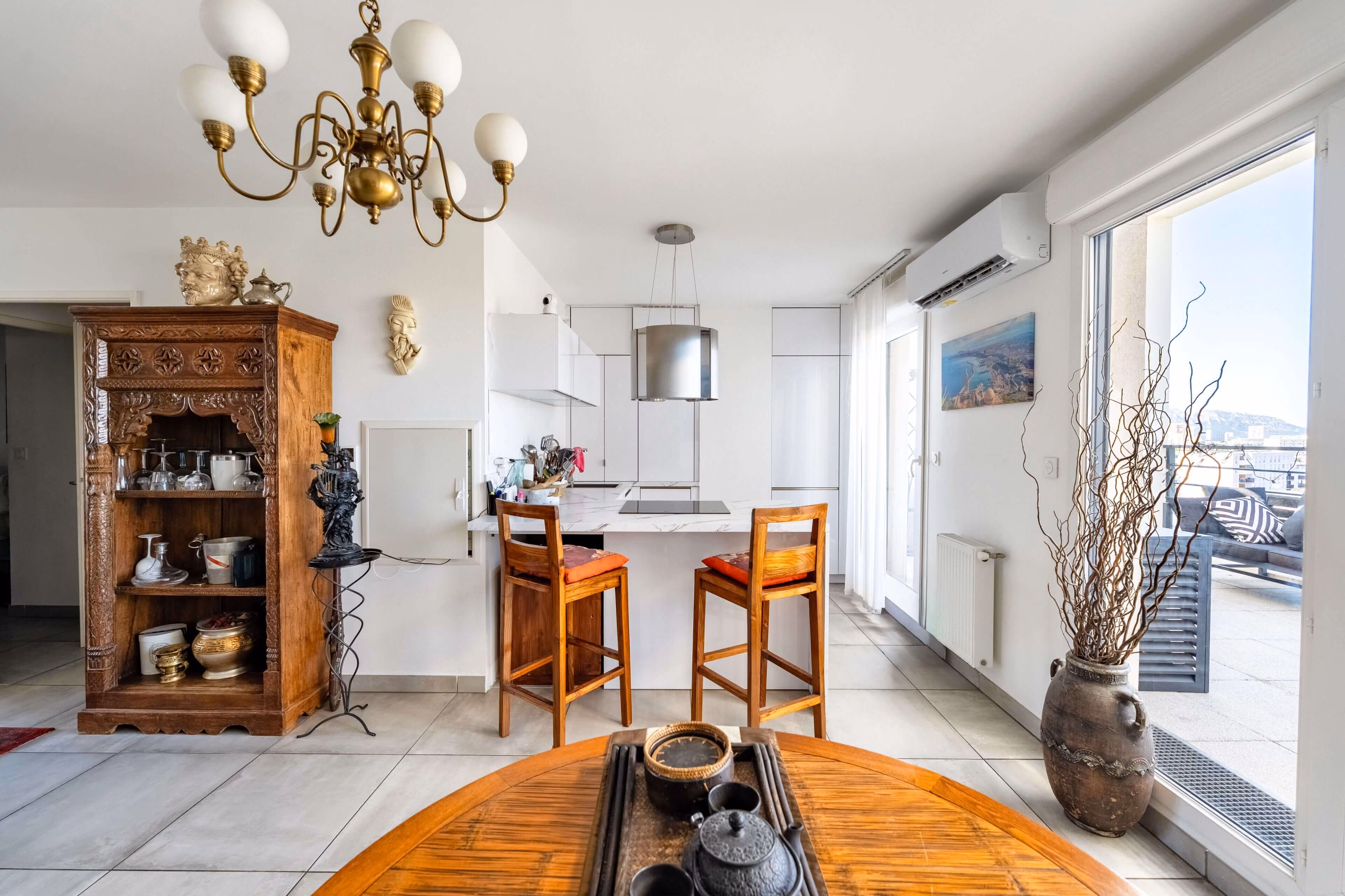 Cuisine moderne blanche avec comptoir, tabourets en bois et balcon avec vue urbaine.
