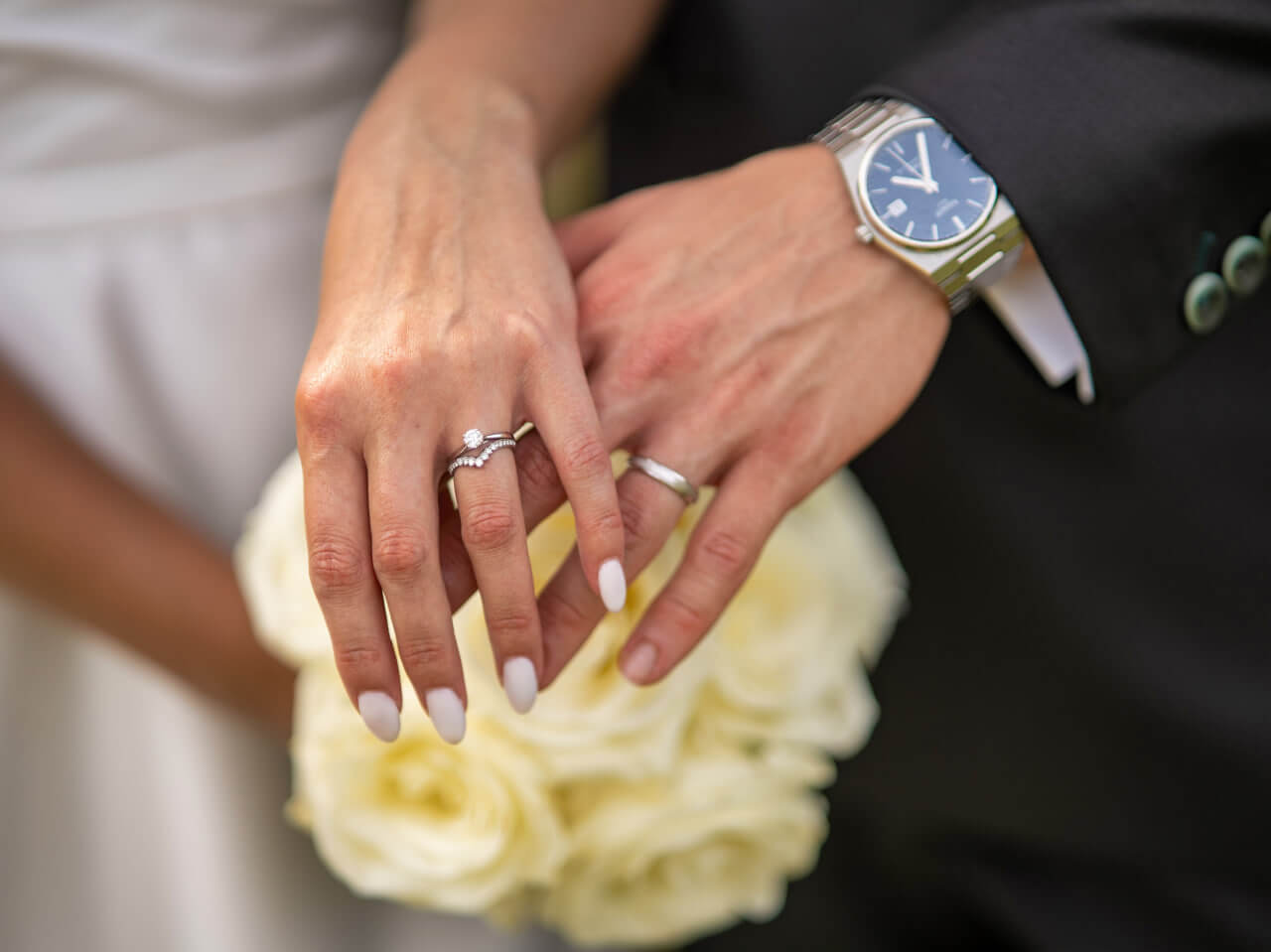 Mains d’un couple avec alliances, femme tenant bouquet de roses blanches, marié en costume noir avec montre.
