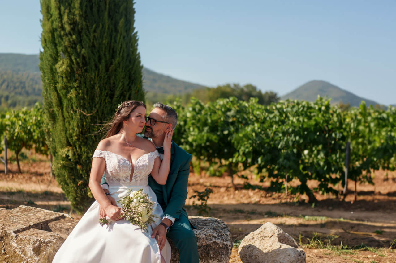 Un couple de mariés assis sur une pierre devant une vigne avec montagnes en arrière-plan.