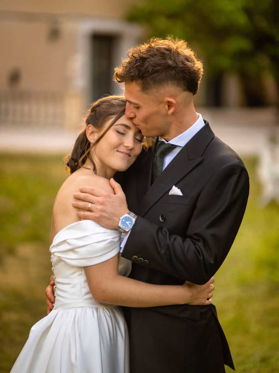 Un couple en tenue de mariage s'embrasse tendrement en extérieur avec de la verdure en arrière-plan.