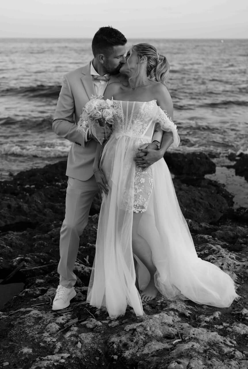 Un couple de mariés s'embrassant sur des rochers près de la mer, la mariée tenant un bouquet.