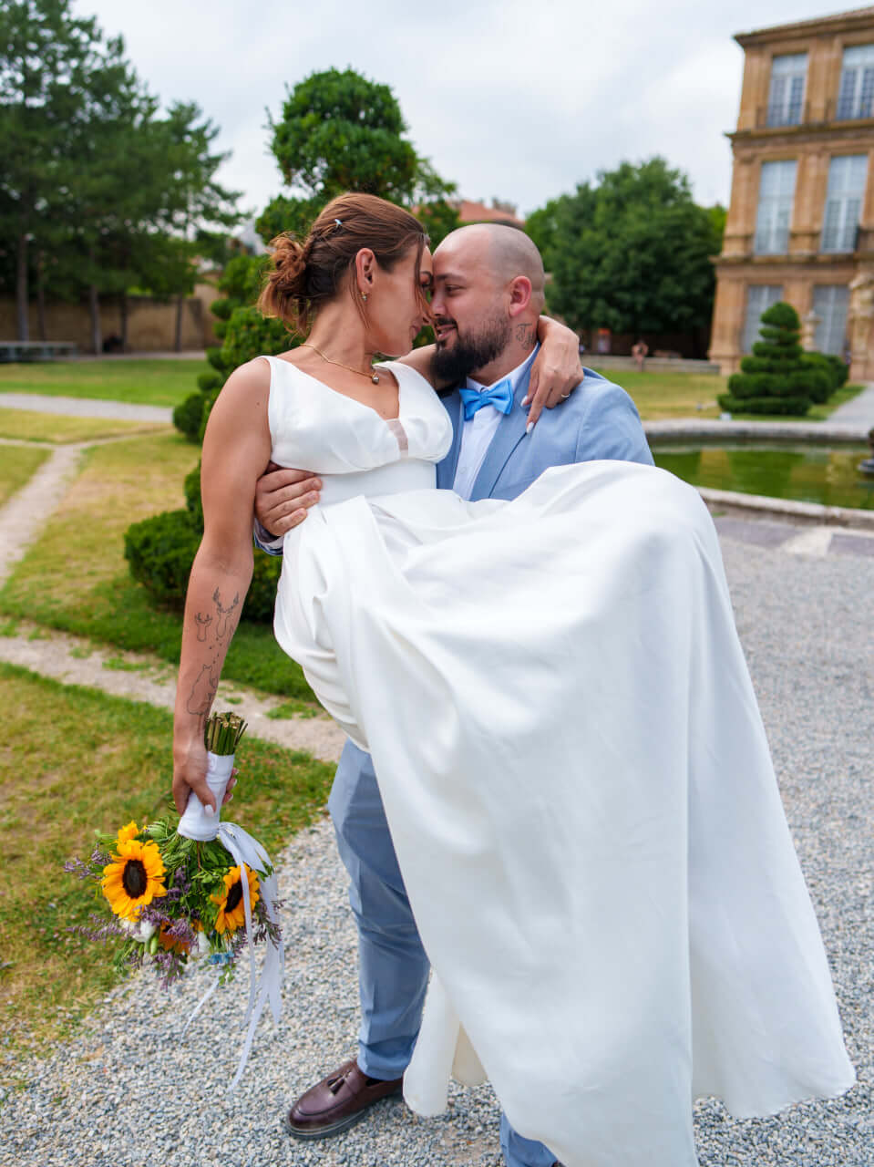 Marié portant sa mariée en robe blanche tenant un bouquet de tournesols dans un jardin.