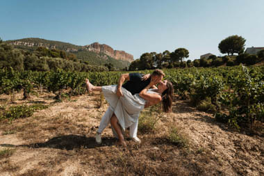 Un homme porte une femme qui rit dans un vignoble avec des montagnes et des arbres en arrière-plan.