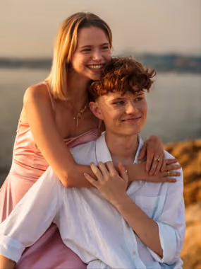 Un jeune couple souriant s'enlace au bord de l'eau au coucher du soleil.