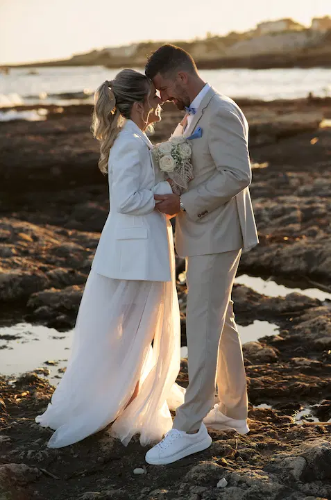Un couple en tenue de mariage se tient face à face sur des rochers au bord de la mer au coucher du soleil.