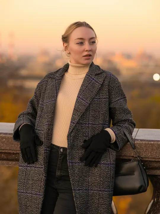 Jeune femme en manteau gris à carreaux, pull col roulé beige, gants noirs, appuyée sur un mur extérieur.
