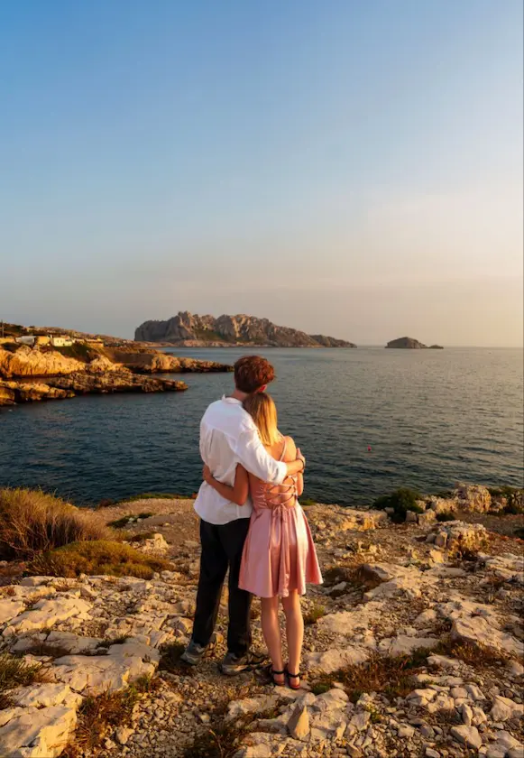 Un couple enlacé admire la mer depuis une roche au coucher du soleil.