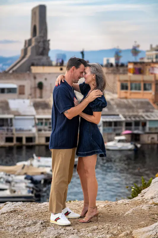 Un couple uni se tient la main sur une plage au coucher du soleil, la femme porte une robe bleue et l'homme une chemise noire.