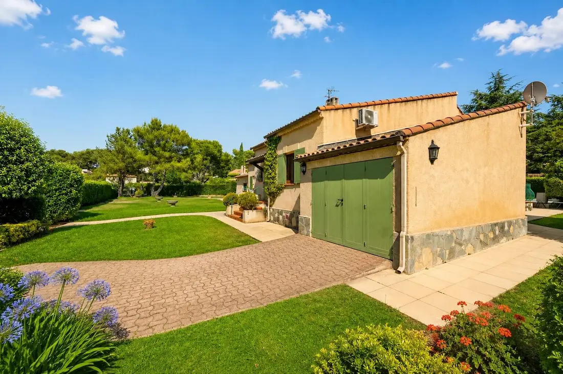Maison beige avec garage vert, allée pavée, jardin verdoyant et fleurs sous un ciel bleu.