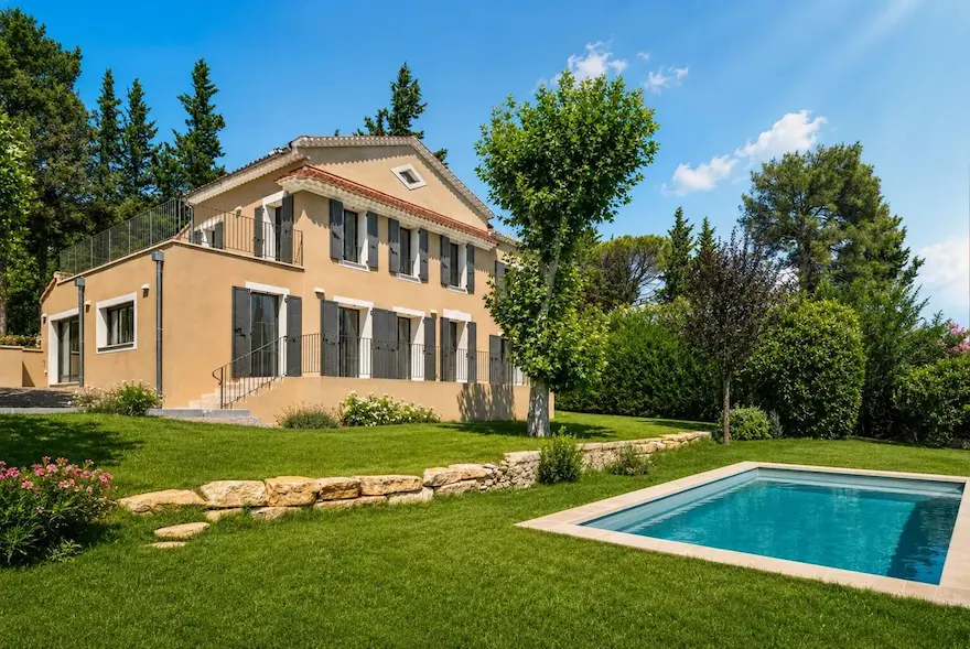 Maison beige avec volets gris, piscine rectangulaire et jardin bien entretenu sous ciel bleu.