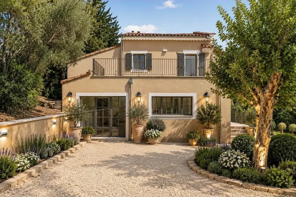 Maison beige avec fenêtres à volets, terrasse, jardin soigné avec arbres et fleurs, allée en gravier.