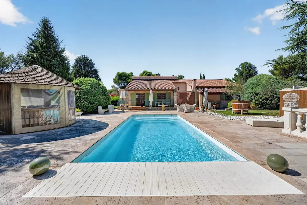 Piscine rectangulaire claire devant une maison entourée de verdure et mobilier extérieur.