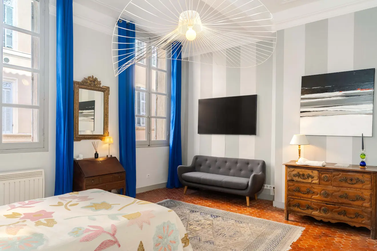 Chambre lumineuse avec lit à couverture fleurie, canapé gris, rideaux bleus, commode en bois et TV murale.