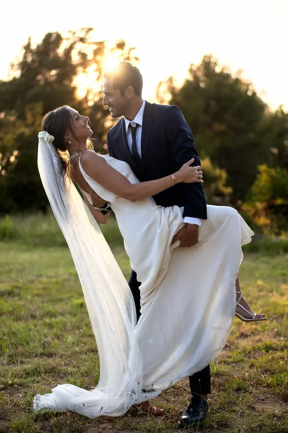 Un homme en costume bleu marine tient une femme en robe blanche de mariée avec voile dans un champ.