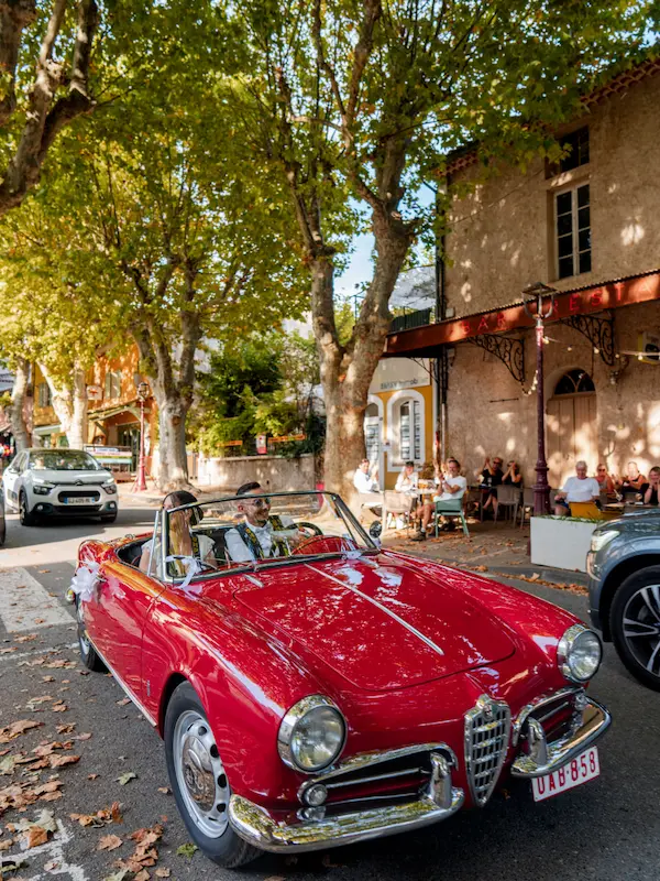 Voiture rouge décapotable vintage conduite dans une rue ombragée bordée d'arbres et de cafés.