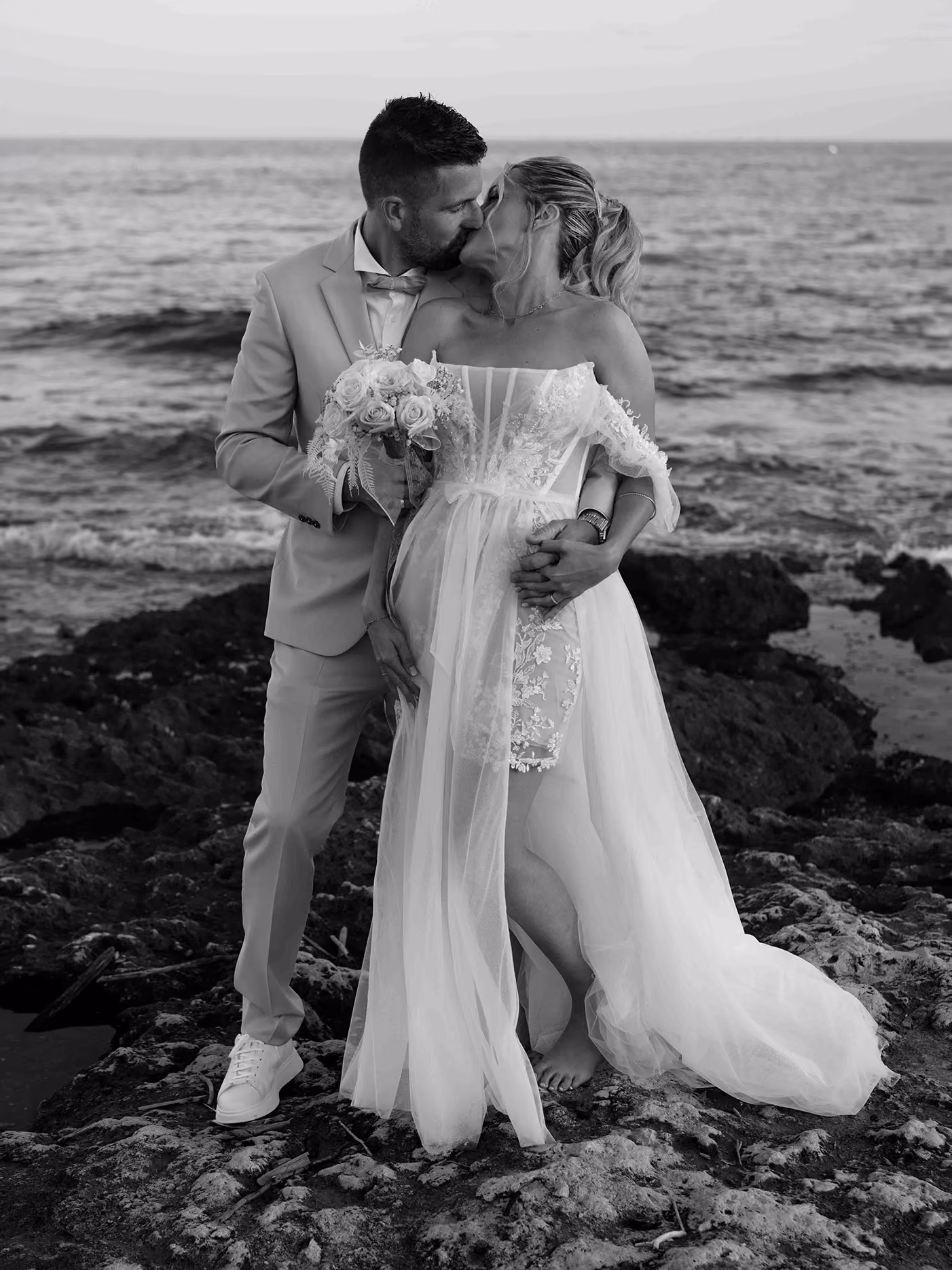 Un couple de mariés s'embrassant sur des rochers près de la mer, la mariée tenant un bouquet.