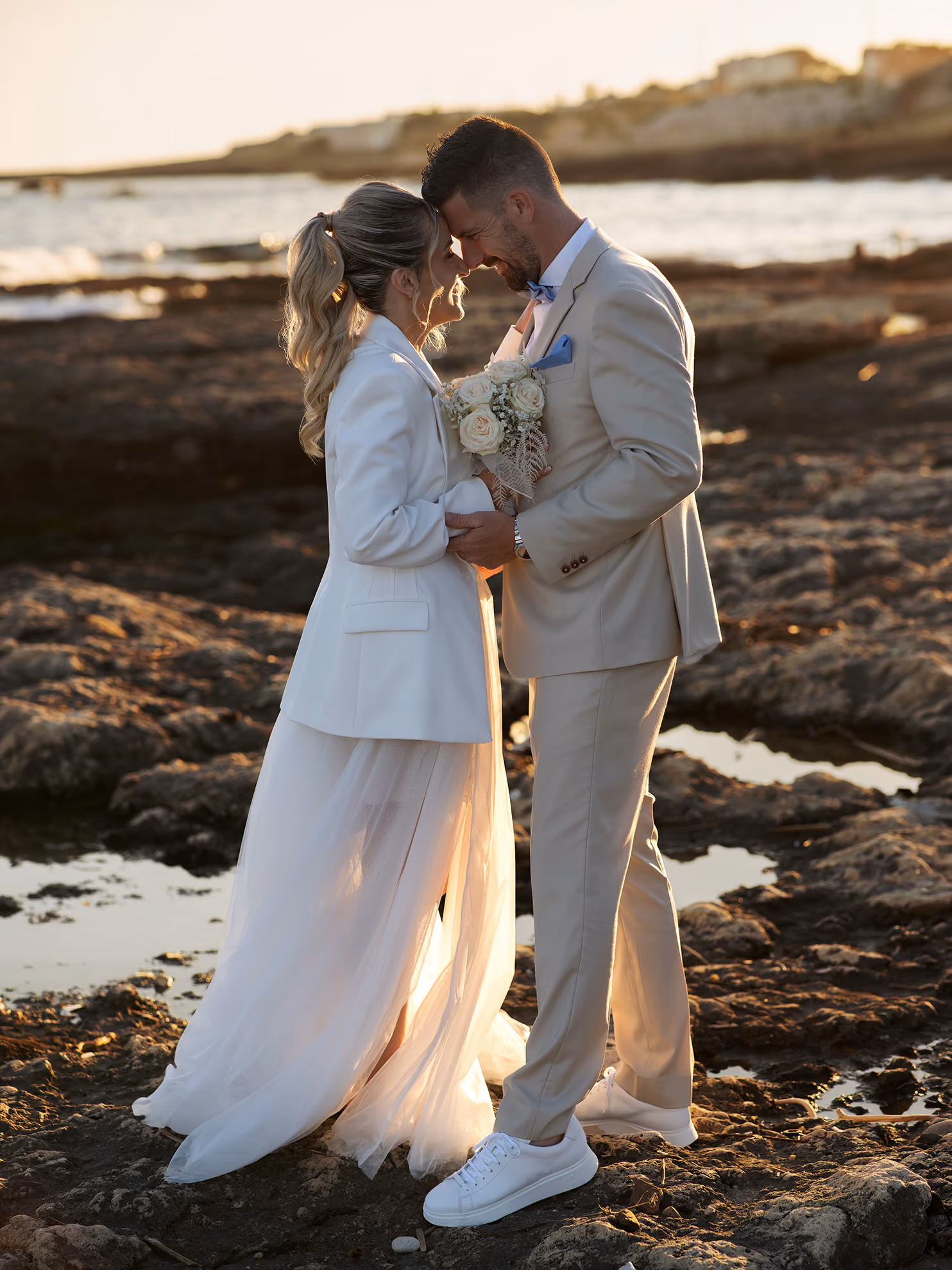 Un couple en tenue de mariage se tient face à face sur des rochers au bord de la mer au coucher du soleil.