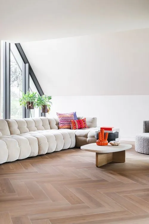 Sala moderna con sofá blanco esquinero, cojines coloridos, mesa de centro redonda y plantas junto a ventanas inclinadas.