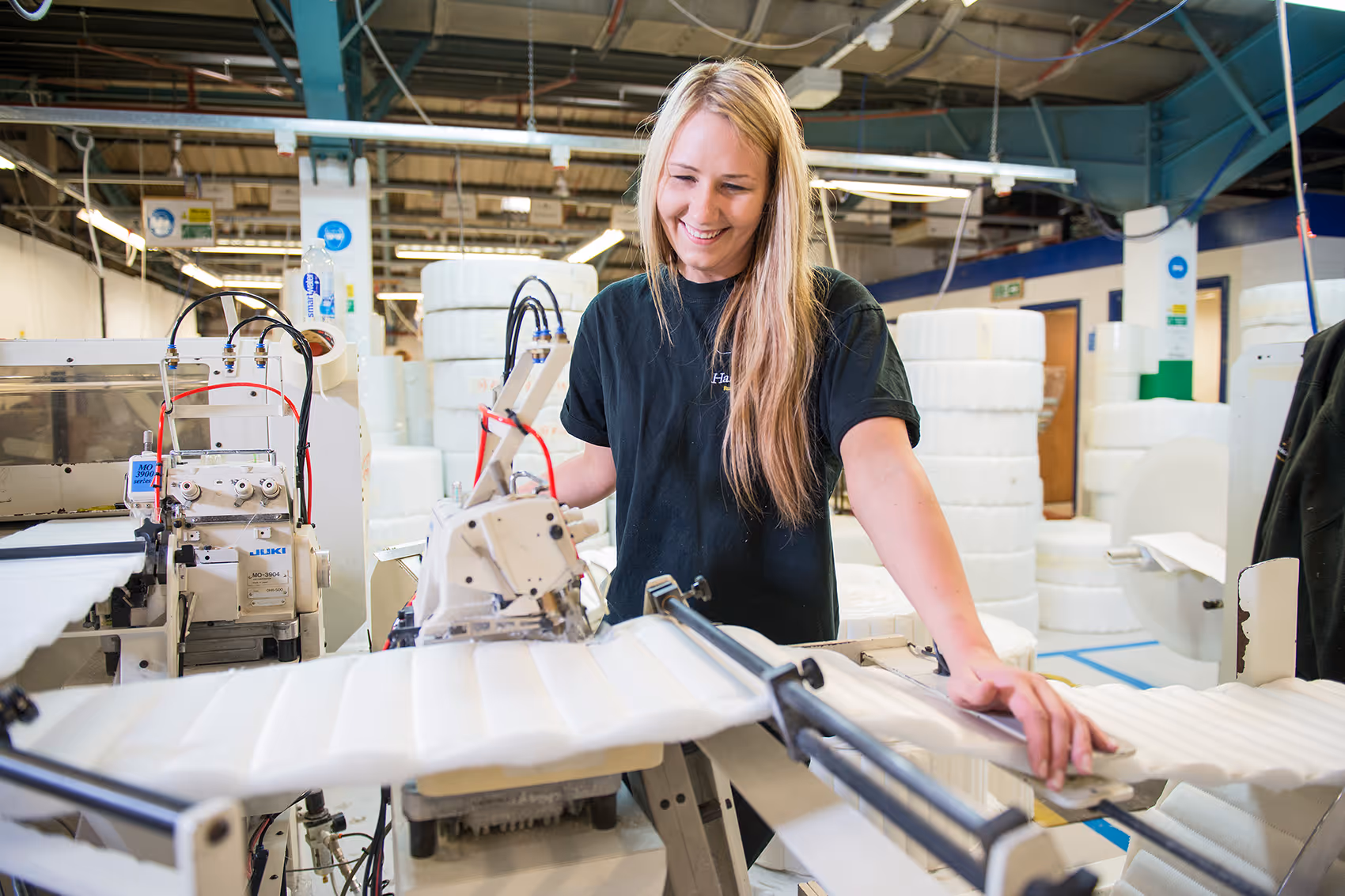 Smiling woman operating a sewing machine in a textile factory with stacked rolls of white fabric in the background.