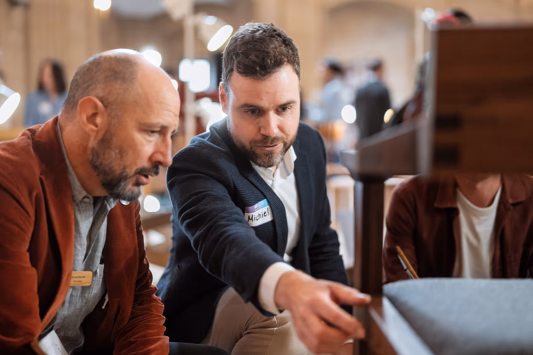 Two men closely examining and discussing a wooden piece of furniture with fabric upholstery in a workshop setting.