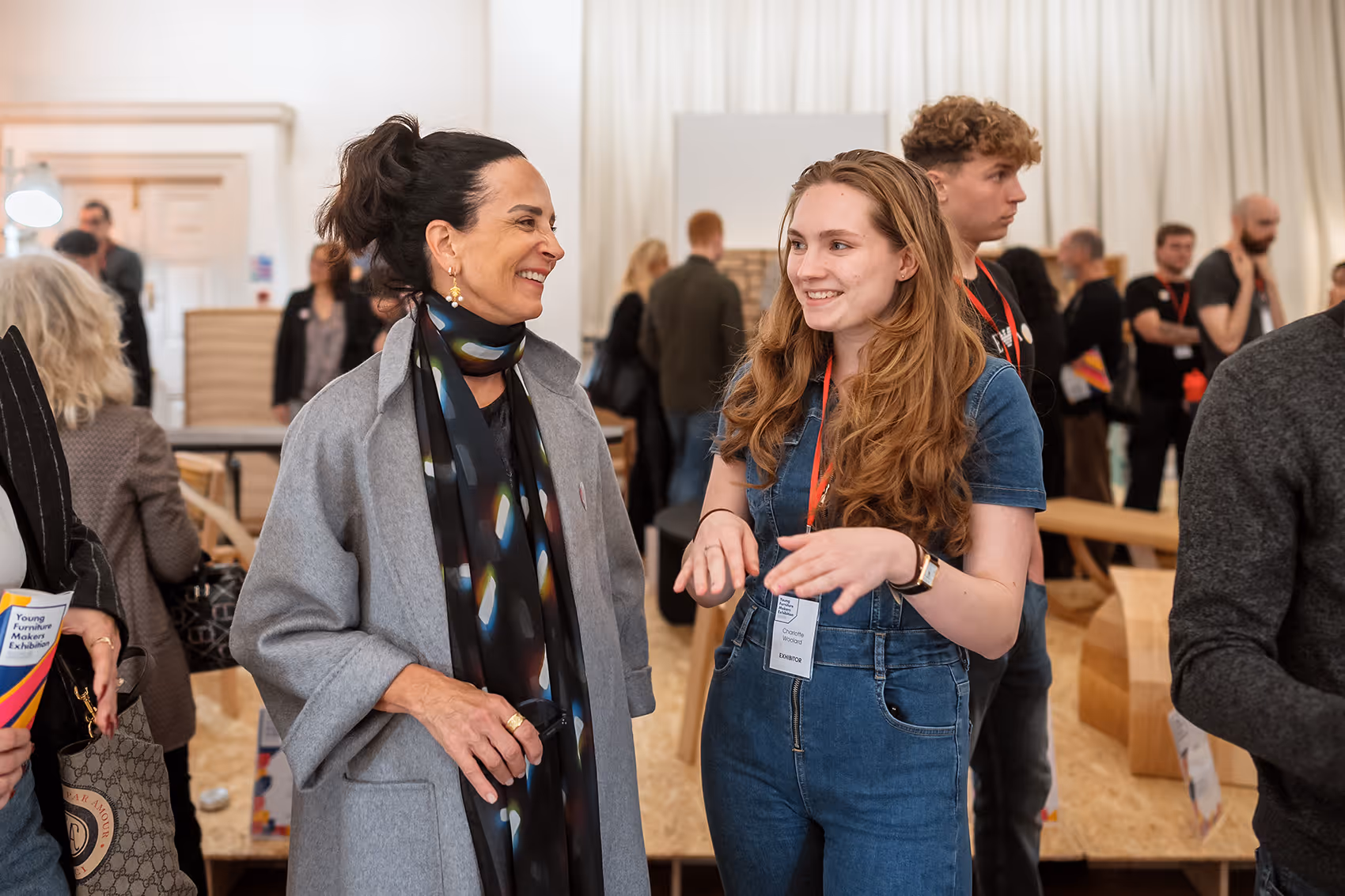 Two women smiling and talking at an indoor event with other attendees in the background.