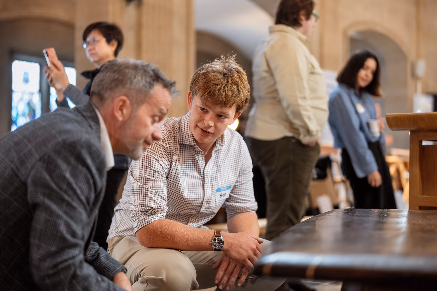 Two men engaged in a conversation, one wearing a checkered shirt with a 'Ben' name tag, with two people standing and interacting in the background inside a spacious room.