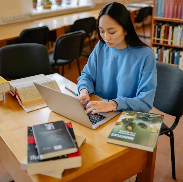 Jeune étudiante asiatique concentrée travaillant sur un ordinateur portable à la bibliothèque, entourée de livres ouverts et fermés