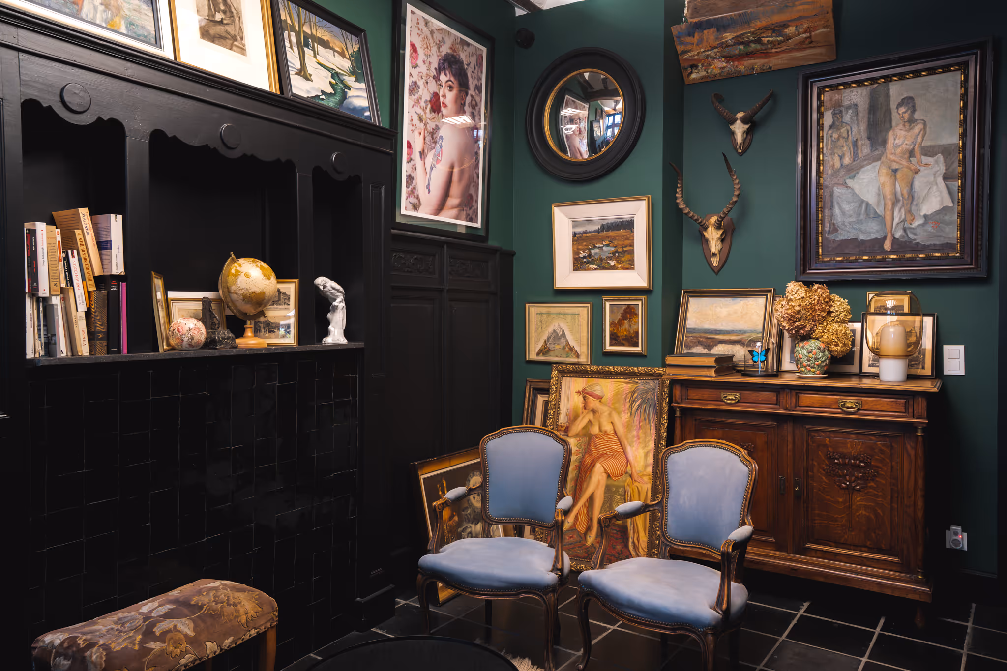 Salon vintage avec deux fauteuils bleus, meubles en bois foncé, diverses peintures sur murs verts, globe terrestre et décorations artistiques.