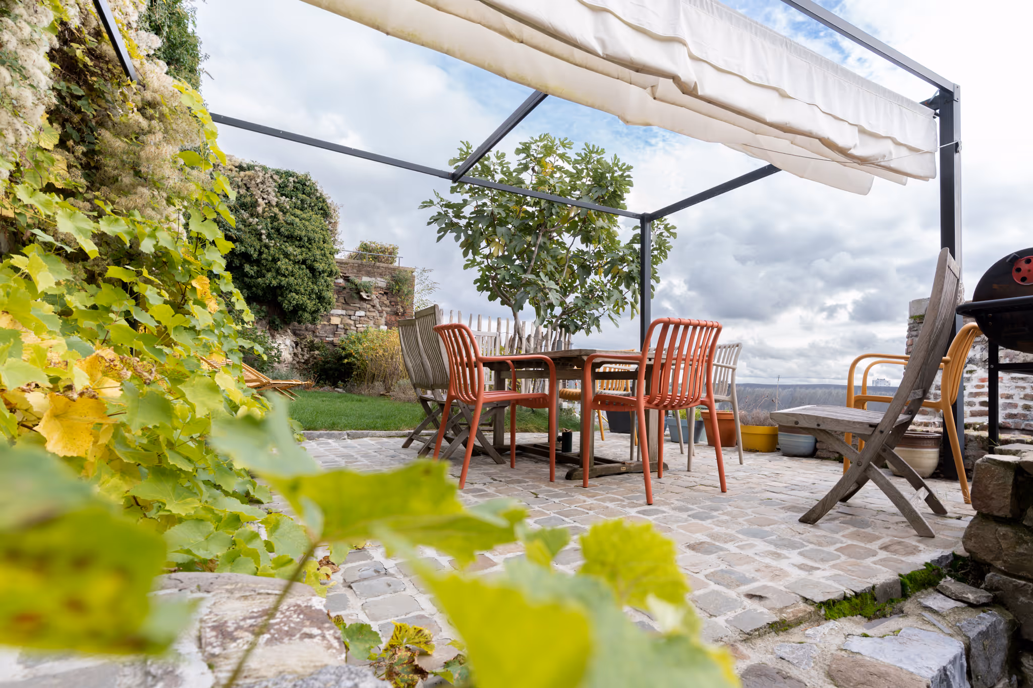 Terrasse en pavés avec une table en bois entourée de chaises colorées sous une pergola avec un store rétractable, et des plantes vertes en arrière-plan.
