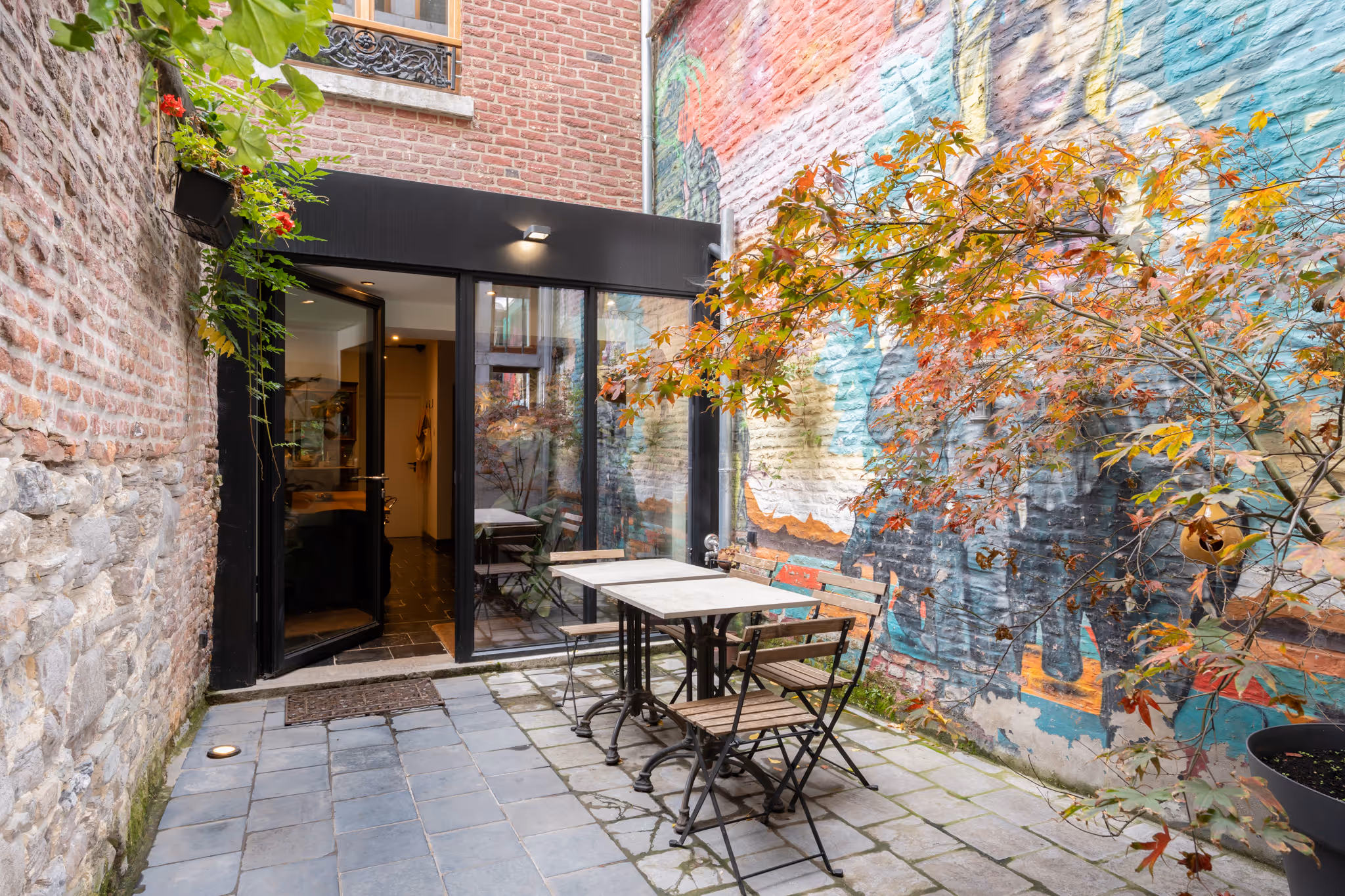 Terrasse extérieure pavée avec une table, des chaises en bois et un mur en briques orné d'une fresque colorée et d'un arbre aux feuilles d'automne.