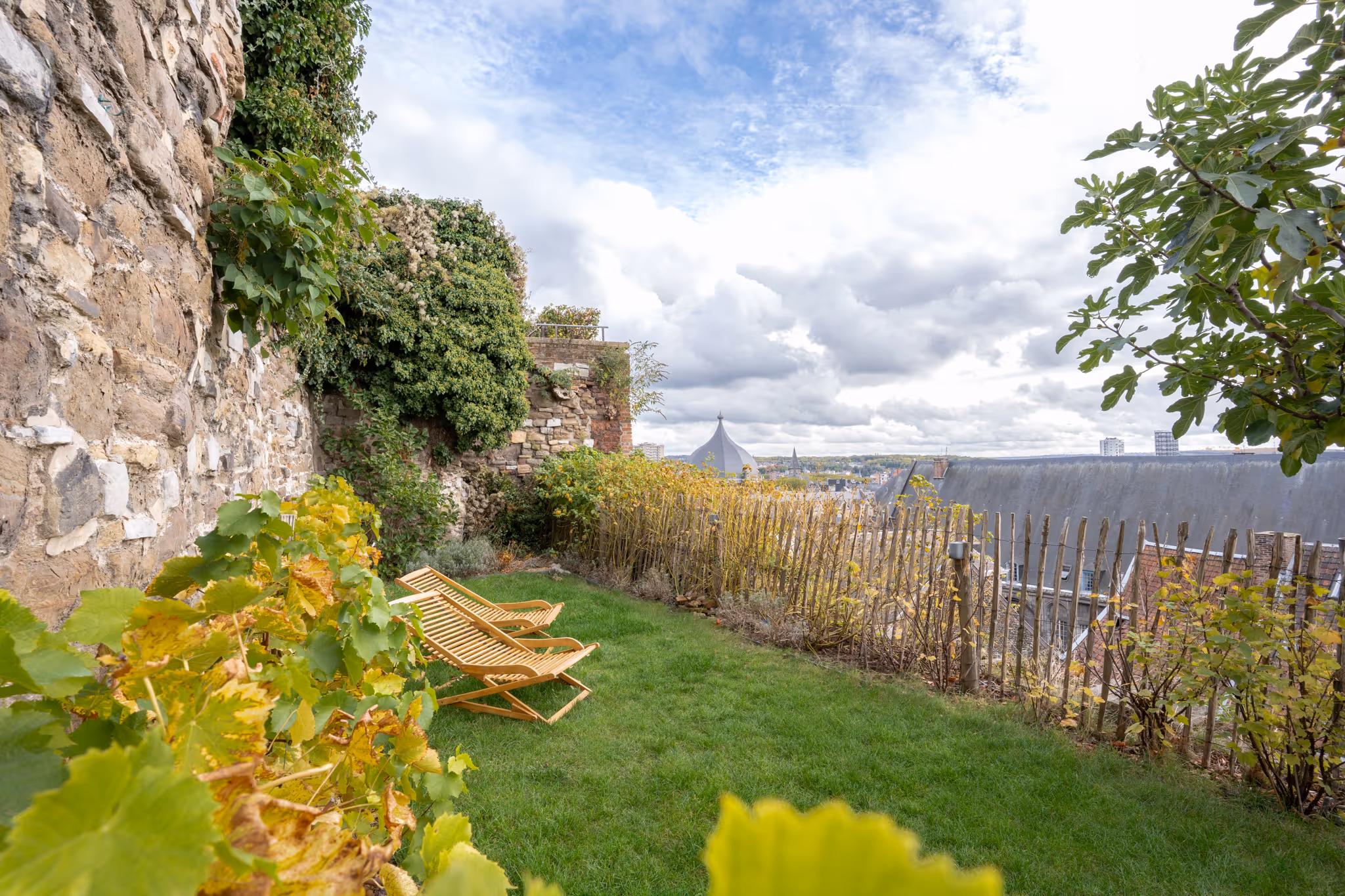 Jardin avec deux chaises longues en bois sur une pelouse près d’un mur en pierre avec vue sur les toits de la ville de Liège
