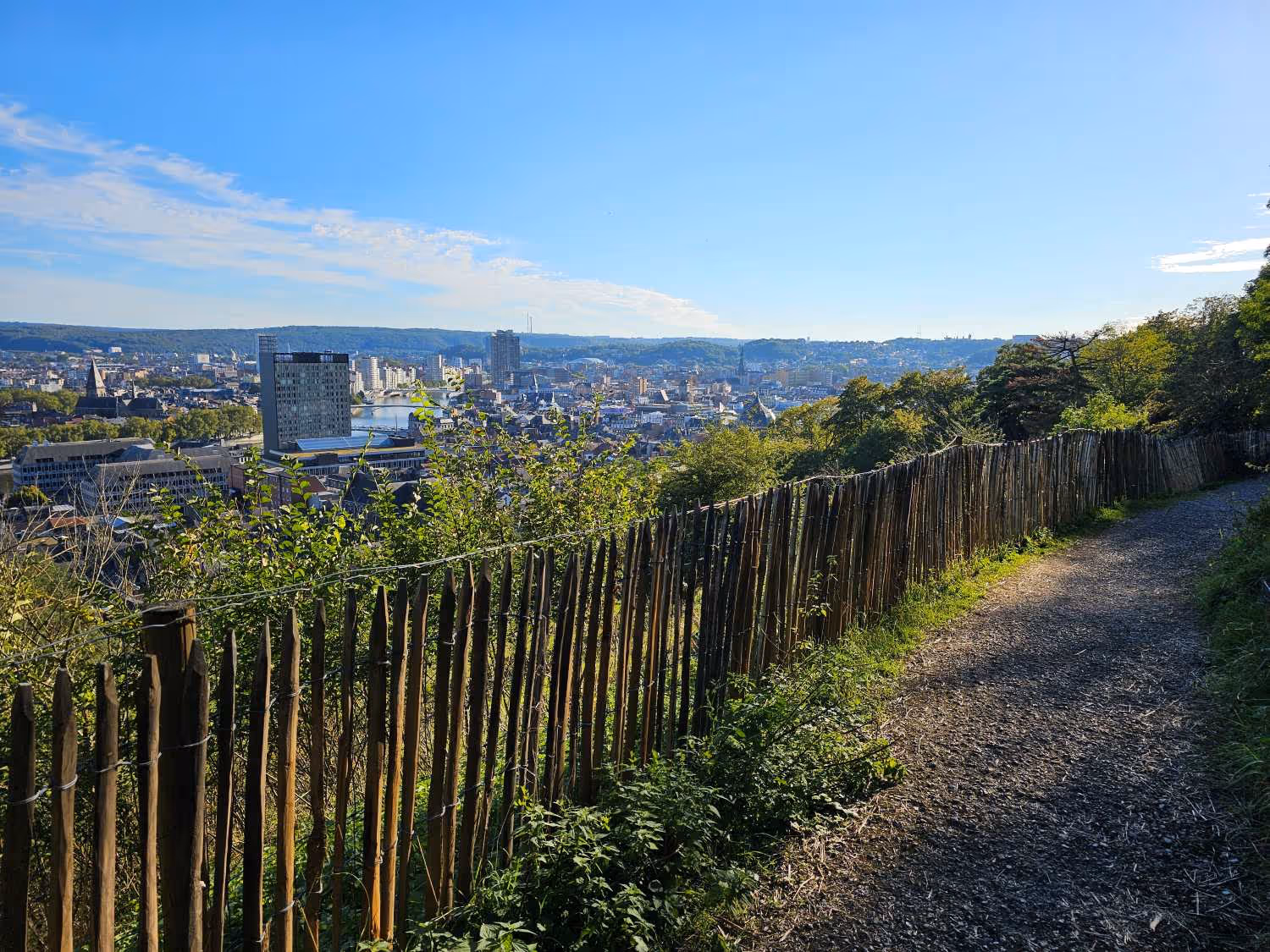 Sentier de randonnée avec vue panoramique sur une ville de Liège sous un ciel bleu clair.