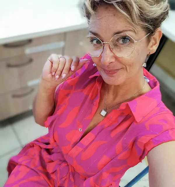 Femme blonde souriante portant des lunettes à monture dorée et une chemise colorée à motifs géométriques roses et rouges, posant pour un selfie en intérieur