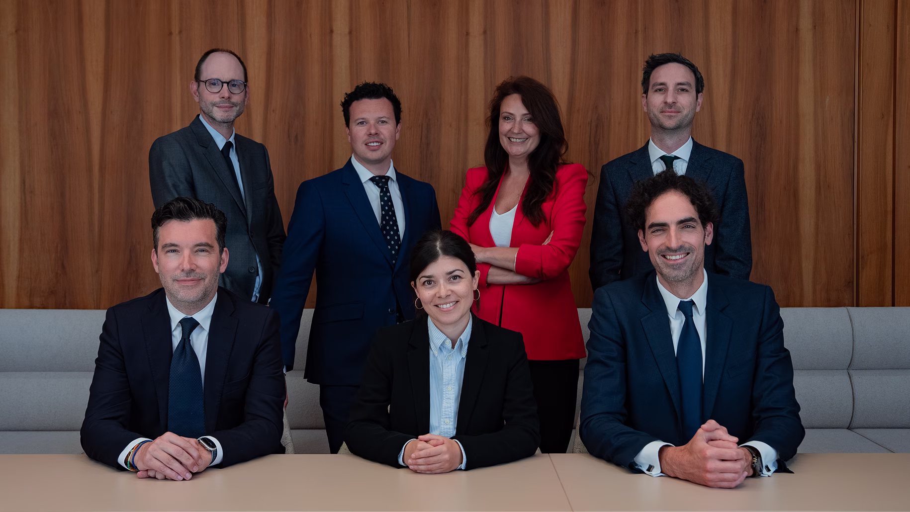 Groupe de sept professionnels vêtus de costumes formels posant devant un mur en bois dans un cadre de bureau.