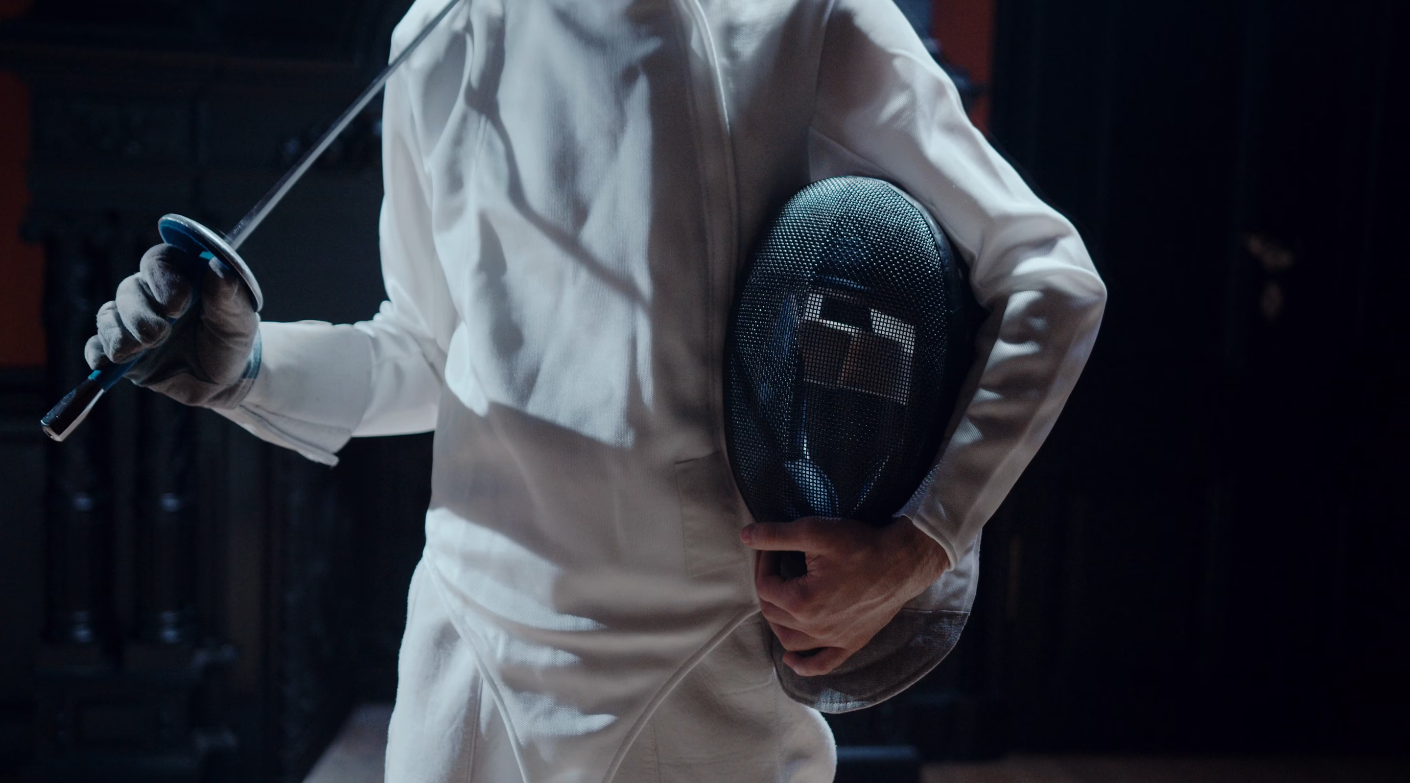 Person en tenue d'escrime blanche tenant un masque d'escrime et une épée.