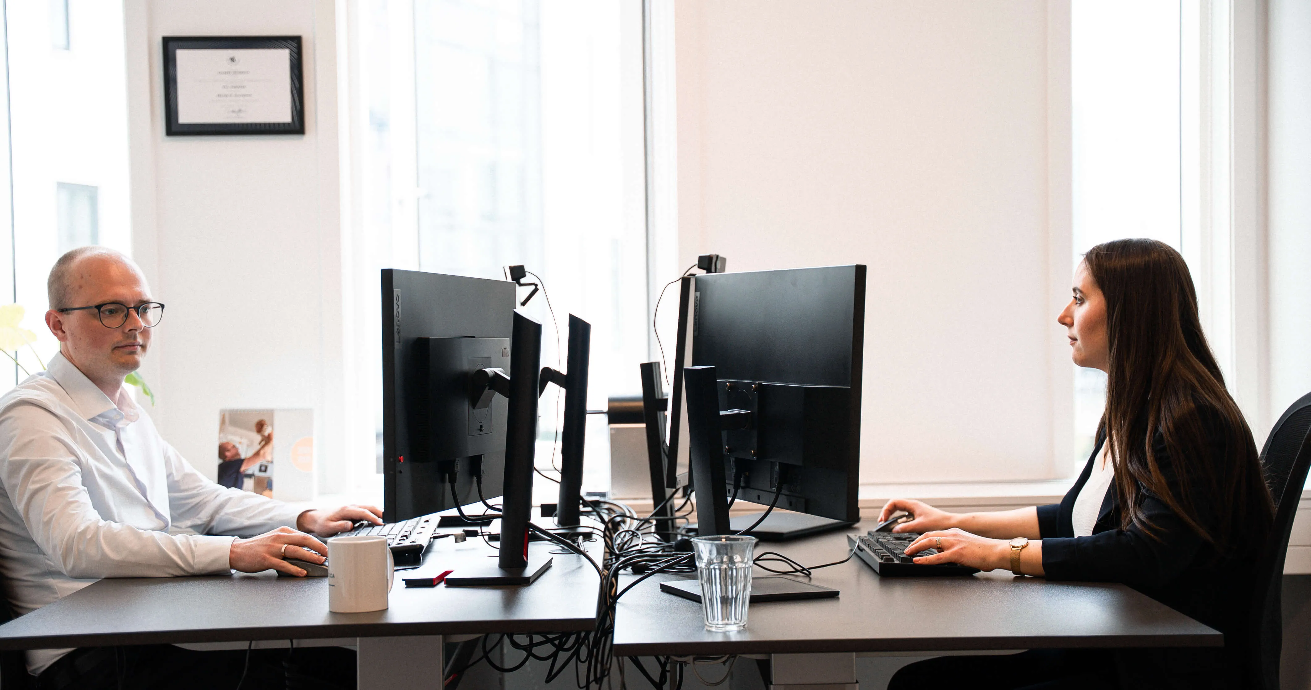 Mand og kvinde sidder overfor hinanden ved hvert deres skrivebord og kigger ind i hver deres computerskærm