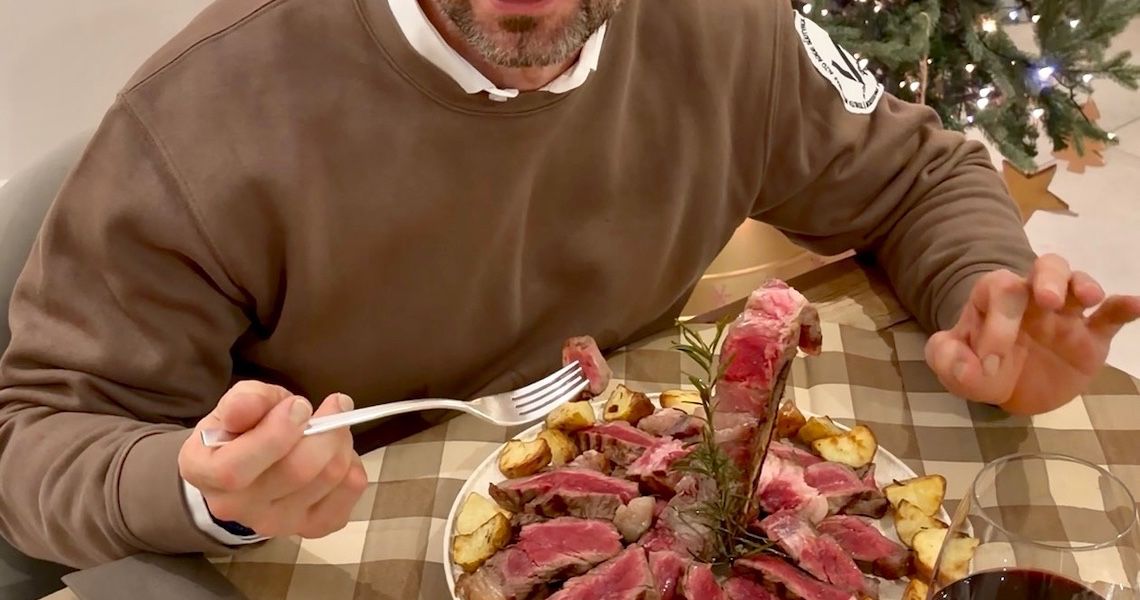 Uomo con occhiali neri seduto a tavola davanti a un piatto con bistecca tagliata e patate arrosto, con un bicchiere di vino rosso e un albero di Natale sullo sfondo.
