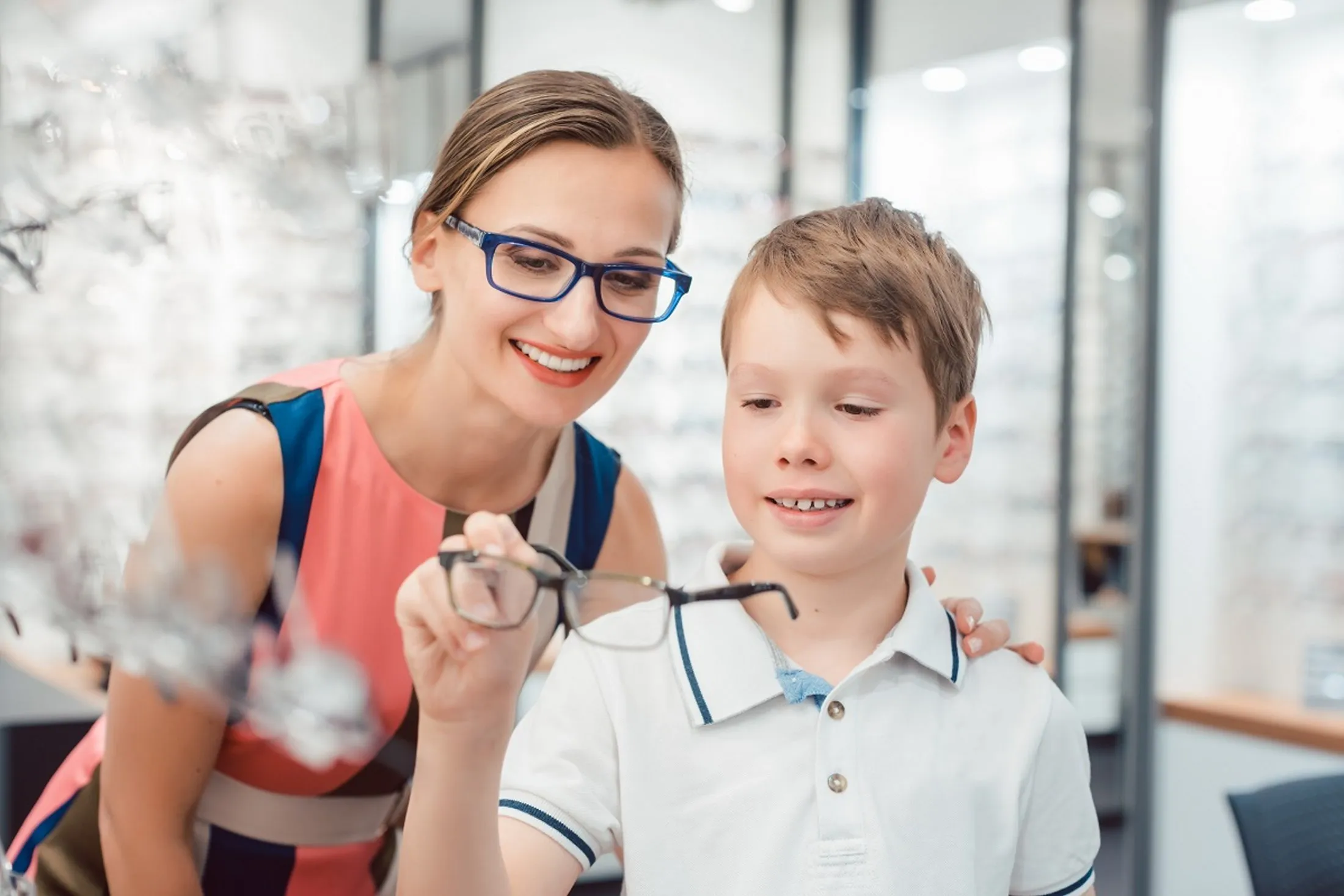 Smiling woman helps young boy try on glasses in optical store