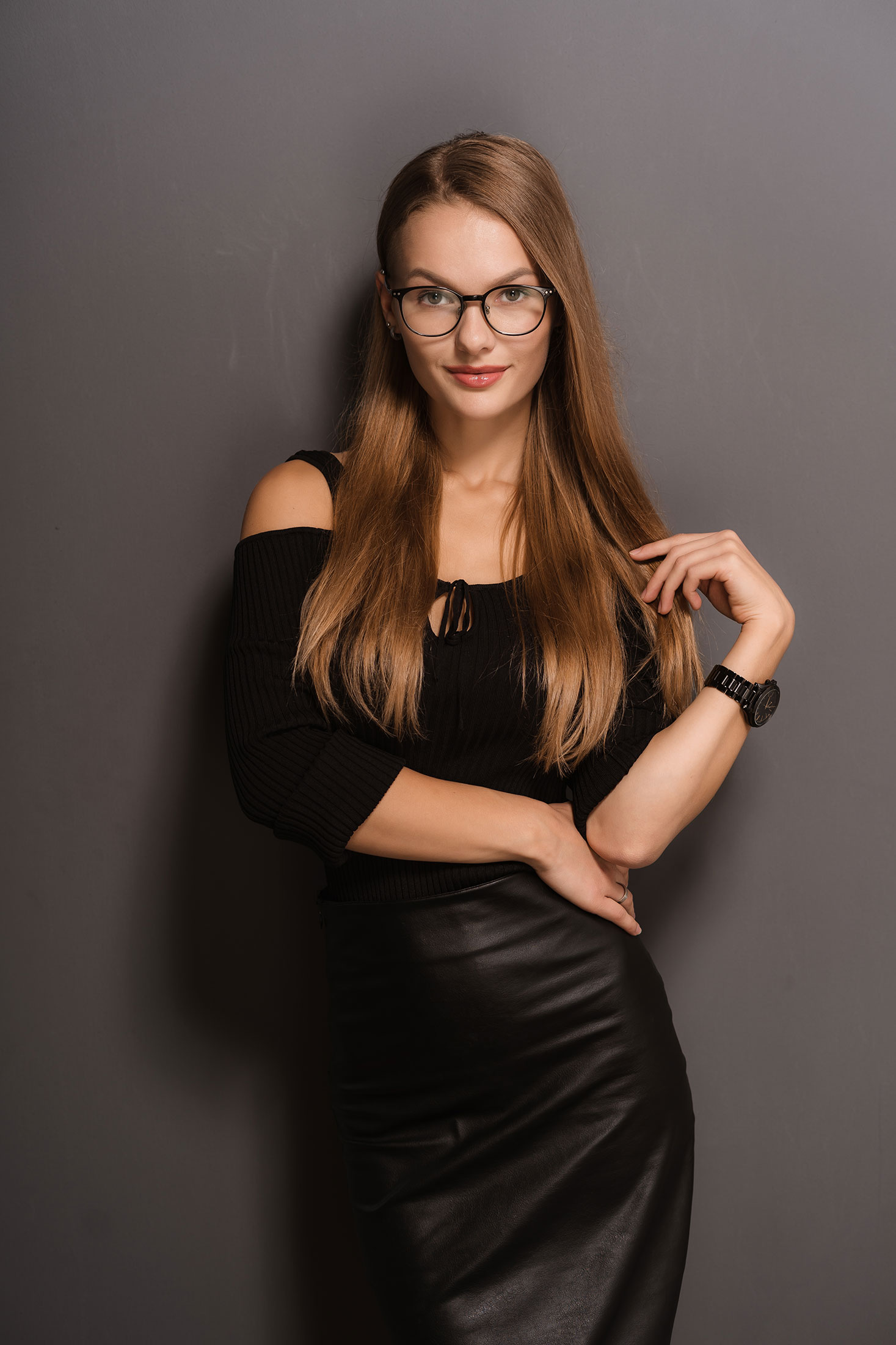 Woman in black off-shoulder top and leather skirt posing against gray wall