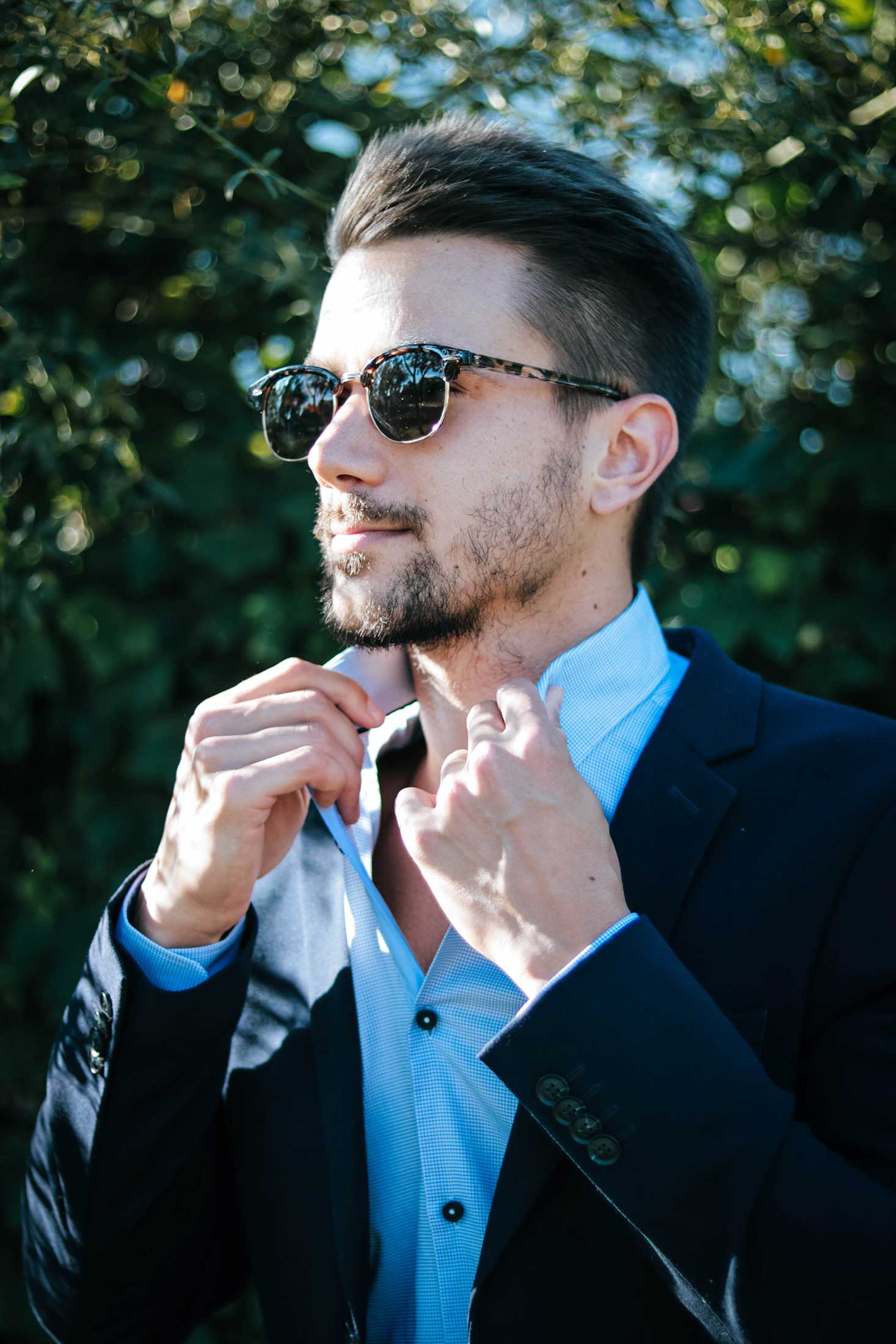 Person in sunglasses adjusting collar, blue shirt and navy suit, leafy background