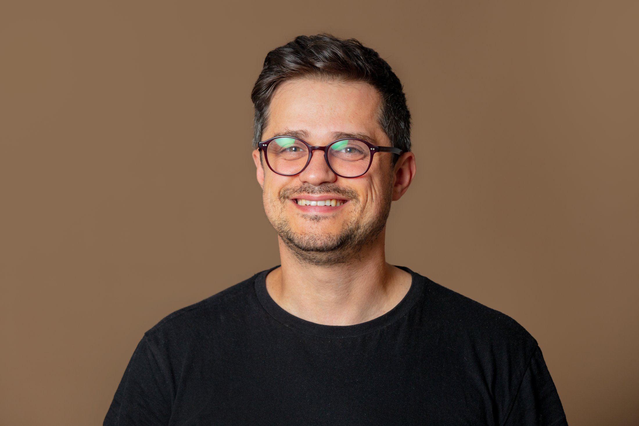 Smiling person with glasses wearing black shirt against brown background