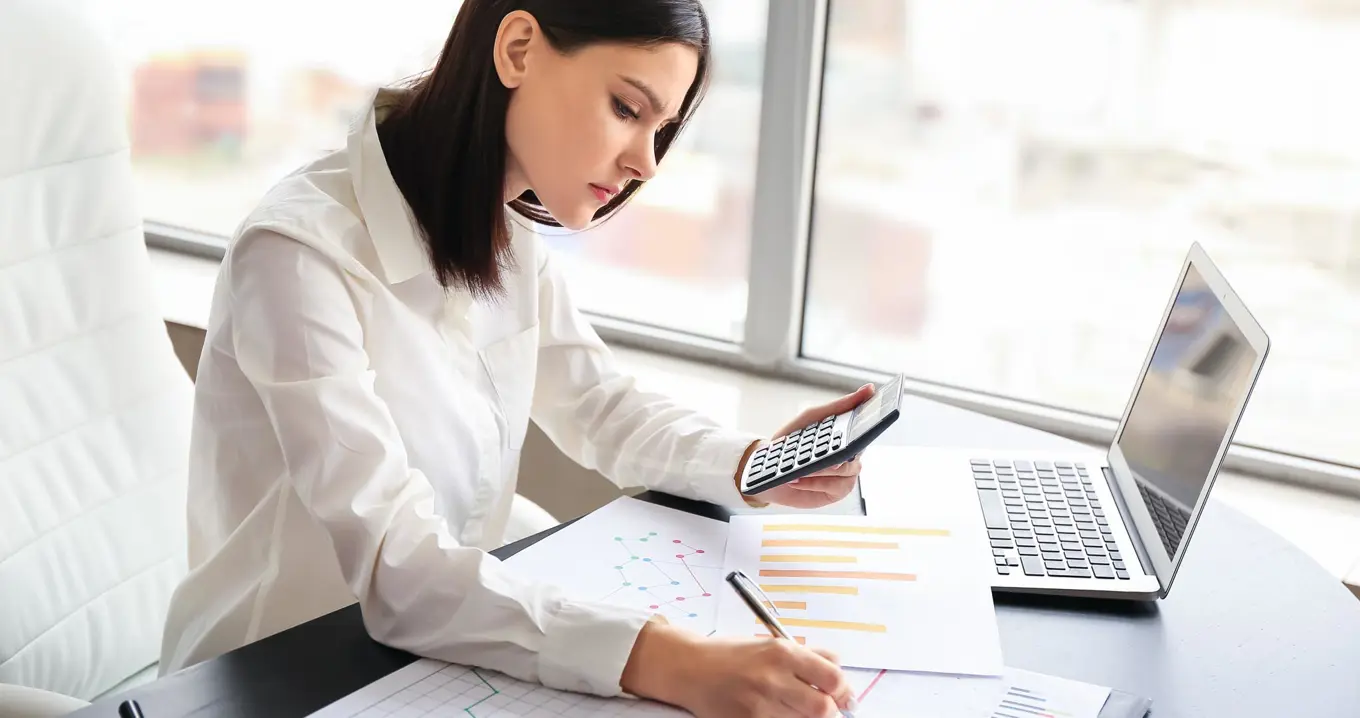 Focused financial controller analyzing graphs and budget data at a desk, using a calculator and laptop to prepare financial reports and forecasts.