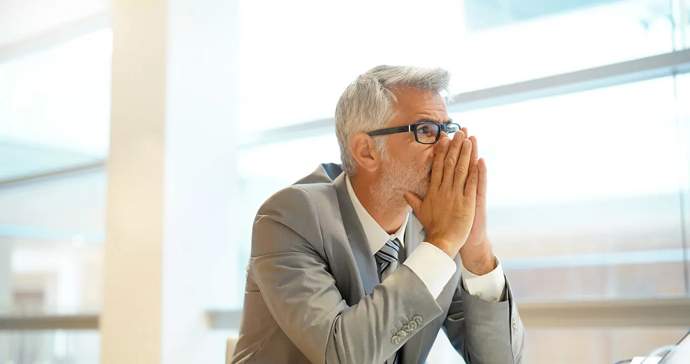 Senior businessman in suit sitting at desk, reflecting on risk management responsibilities and potential business challenges.